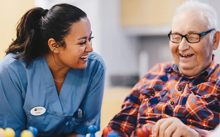 Kvinna i blå vård-arbetskläder skrattar tillsammans med äldre man i rutig skjorta.