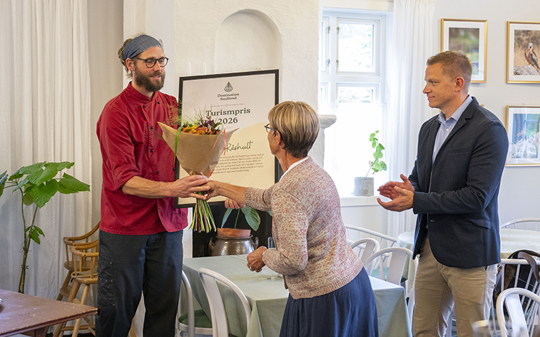Magnus håller ett stort diplom i ena handen och med den andra tar han emot en bukett blommor från Sara. Intill står Erik och applåderar.