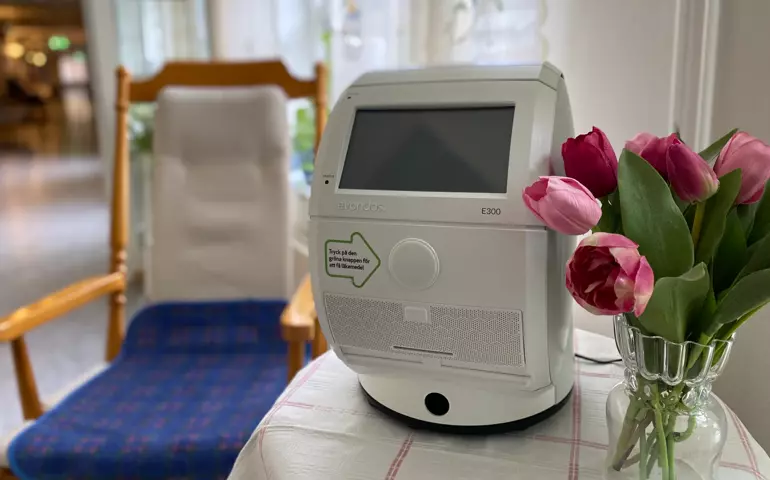 En avlång, lite lådliknande, maskin med ett fönster för digital text står på ett bord med tulpaner. Jämte bordet står en gungstol.