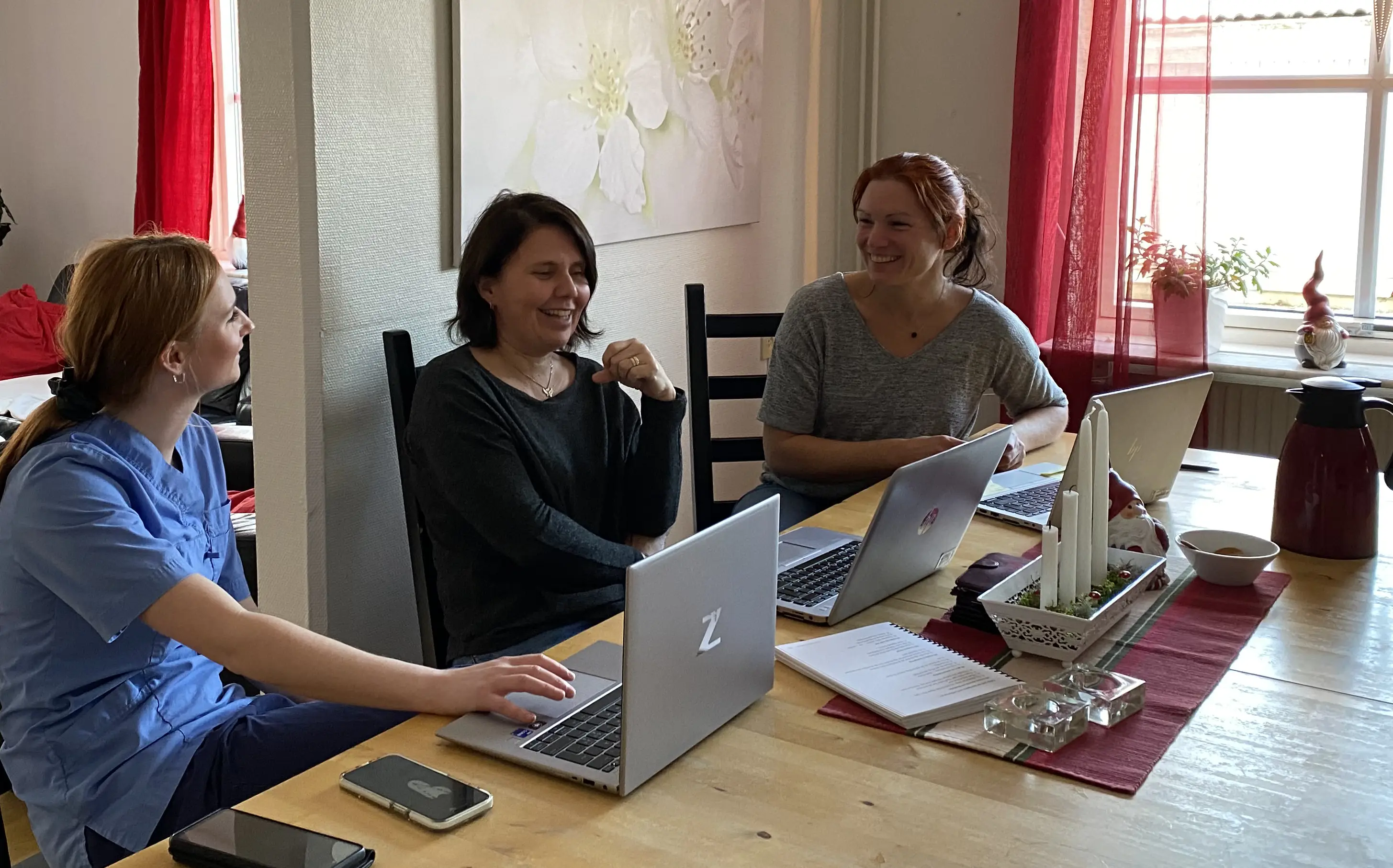 Tre kvinnor sitter vid ett bord i furu, pratar och noterar på sina datorer.