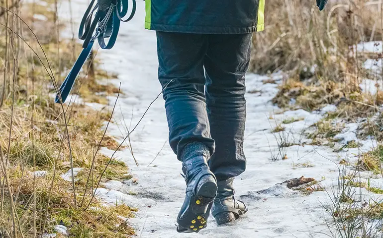 Person som går med broddar och ett hundkoppel i handen på en snöig stig. Man ser sulan med broddar när personen lyfter foten.