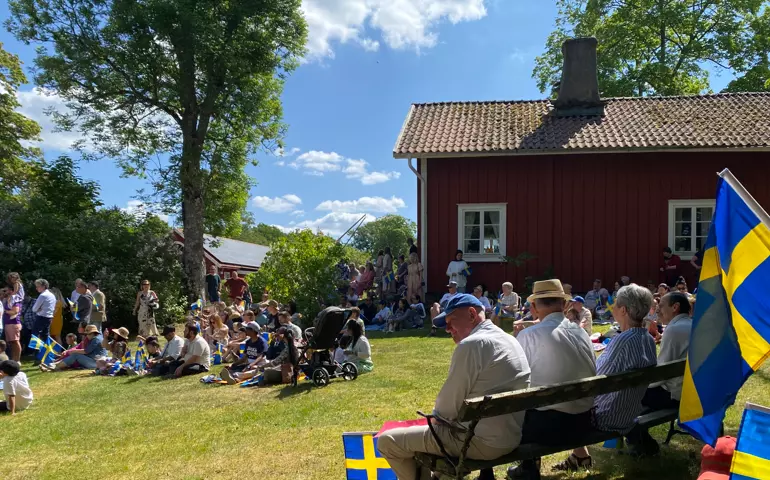 Några personer sitter på en träbänk, några sitter på filtar. De håller i svenska flaggor. Soligt.