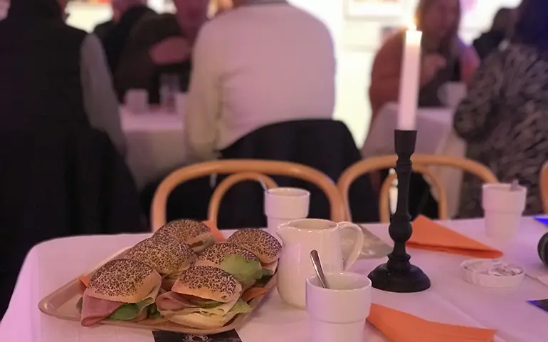 Smörgåsar, kaffekoppar och ett tänt ljus på ett bord med folk i bakgrunden.