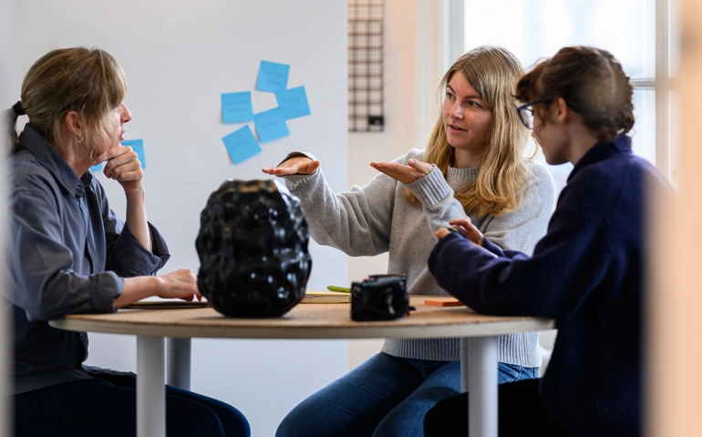 Personer sitter och diskuterar kring ett bord med en vas på. de gestikulerar.