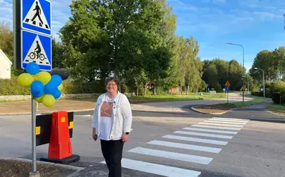 Kvinna står vid ett gång- och cykelöverfartsställe med parkliknande miljö i bakgrunden