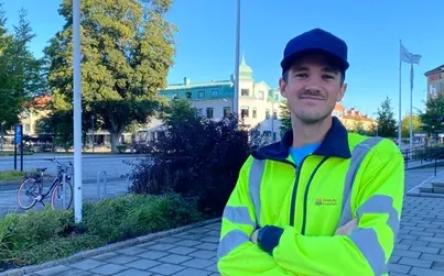 En man står på en trottoar med buskar, träd och hus i bakgrunden. Lite stadsmiljökänsla.