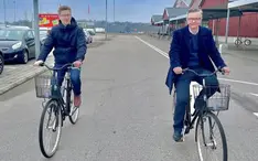 Två mörkklädda män cyklar på varsin cykel vid ett handelsområde
